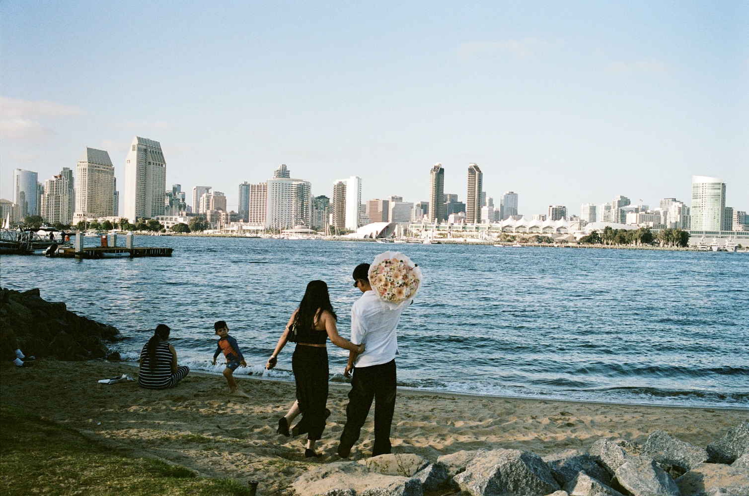 A man carries a large bouquet of flowers across the sand while his partner clutches onto him. The San Diego skyline is in the background.