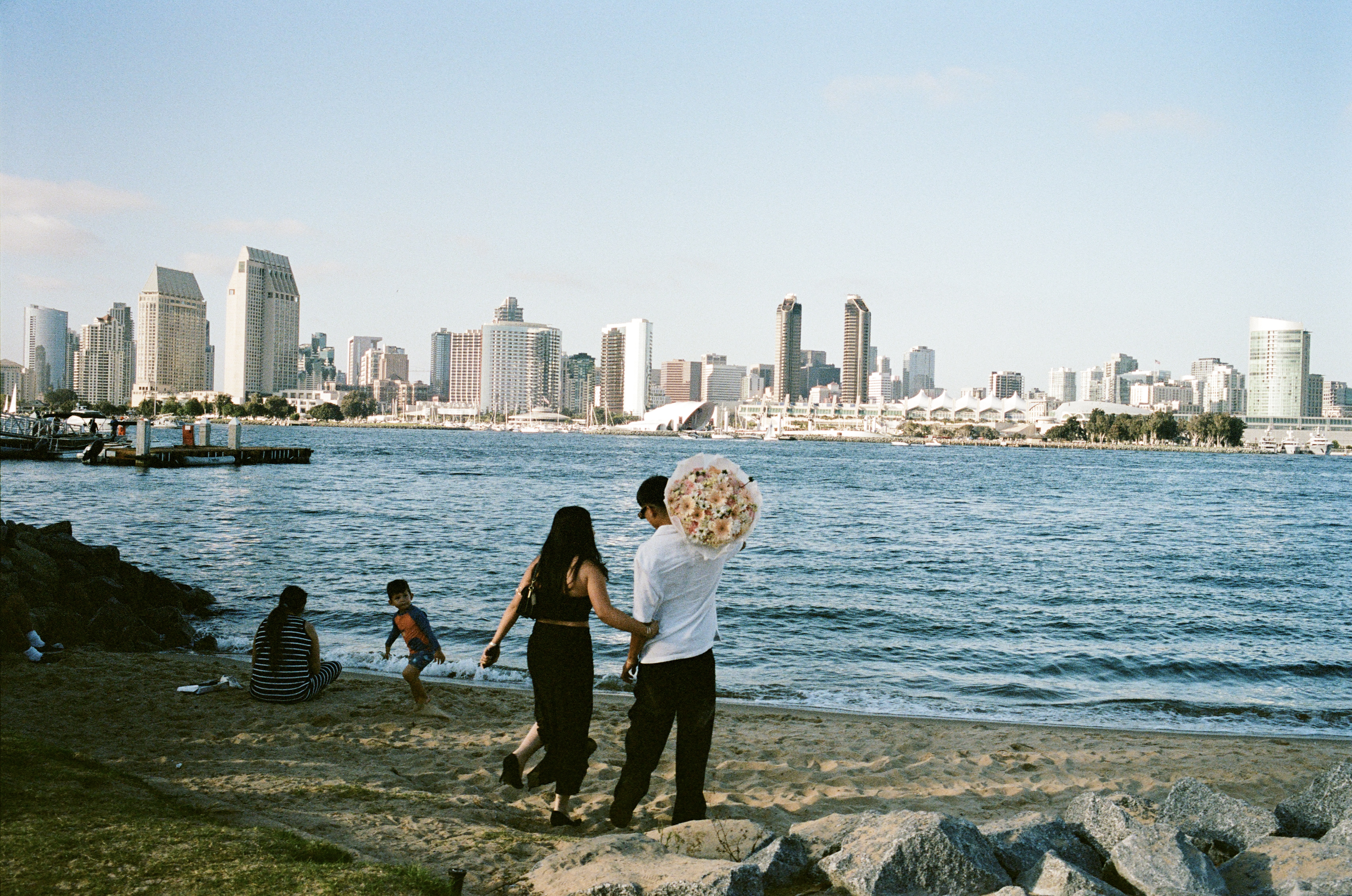 A man carries a large bouquet of flowers across the sand while his partner clutches onto him. The San Diego skyline is in the background.