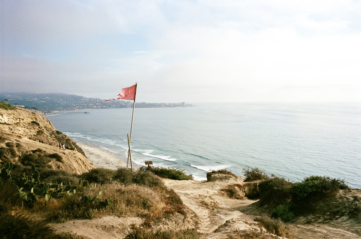 A torn and tattered red flag, wind whipped, sits at the edge of a cliff with La Jolla, California in the background.