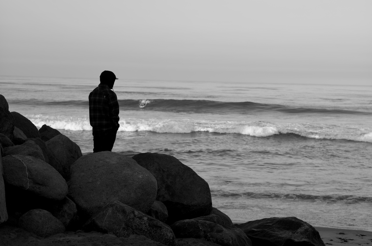 A photograph in black and white shows a man in a hoodie looking out towards a breaking wave from the rocks. In the distance, a surfer is getting ready to ride the wave.