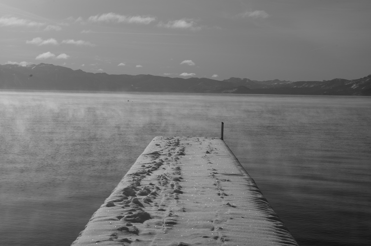 A black and white photograph shows a snow-dusted pier over a lake with mountains in the background. A fog sits over the water, and the snow is sparkling.