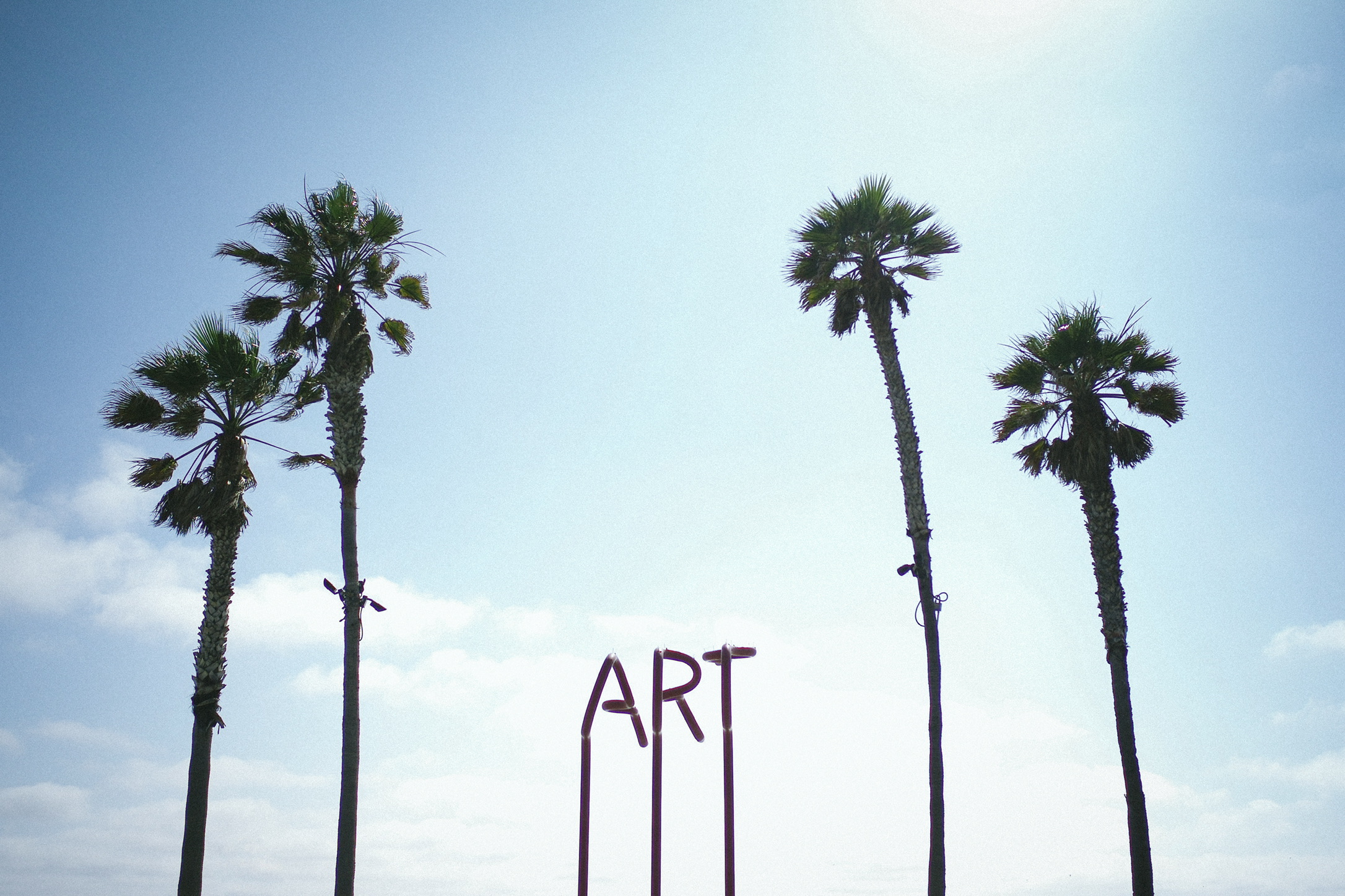 An art installation reads ART and is flanked by two palm trees on both sides, set against a blue sky with clouds.